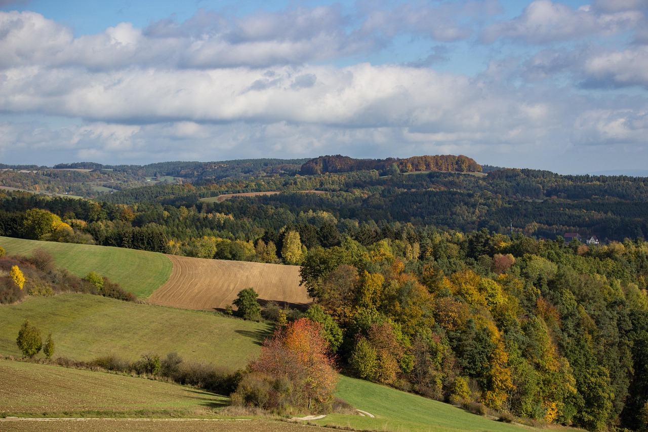 Hügelige Landschaft mit herbstlich gefärbtem Mischwald, Wiesen und abgeernteten Feldern unter teils bewölktem Himmel.