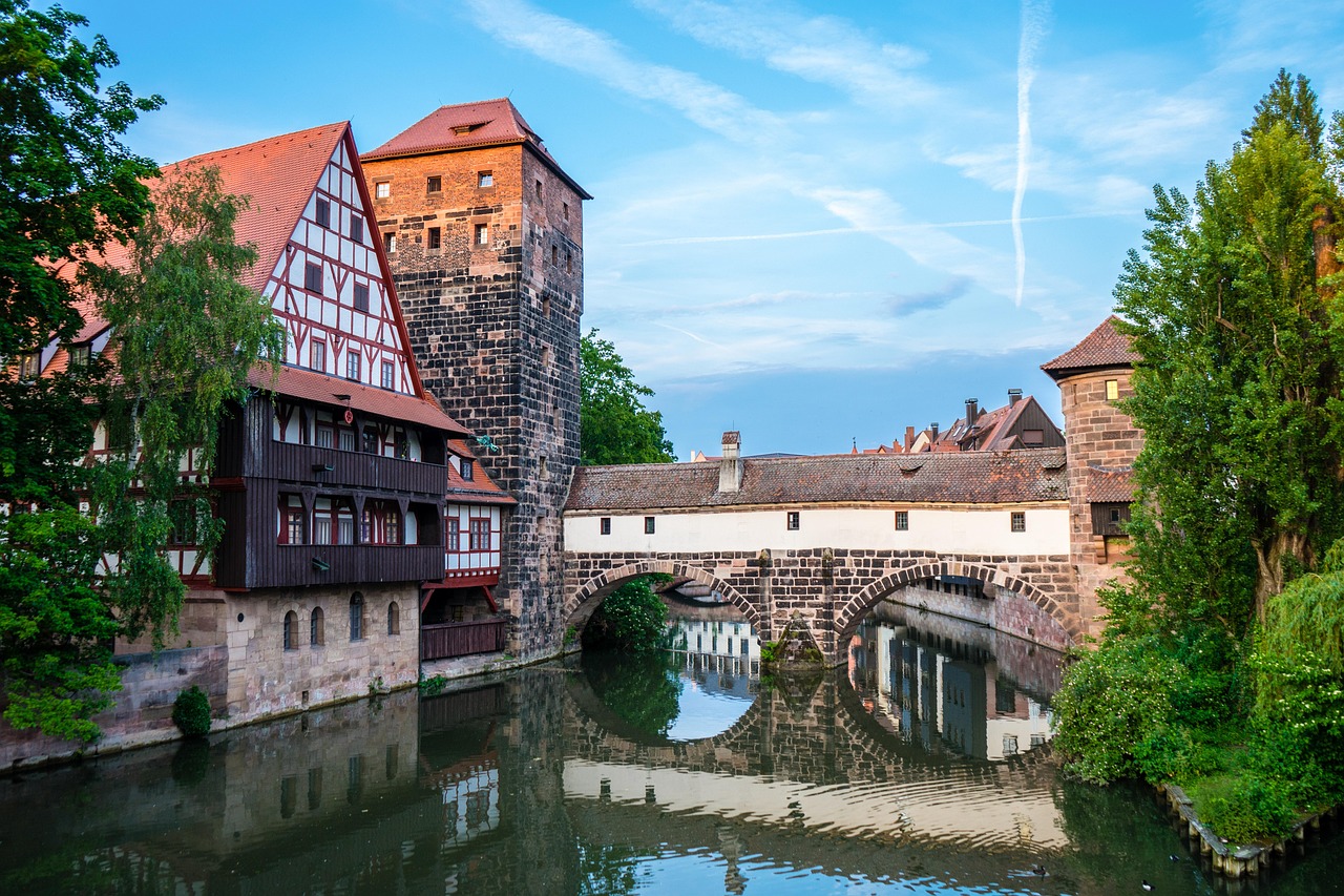 Fachwerkhaus, Wehrturm und überdachte Steinbrücke spiegeln sich im ruhigen Wasser eines Flusses.