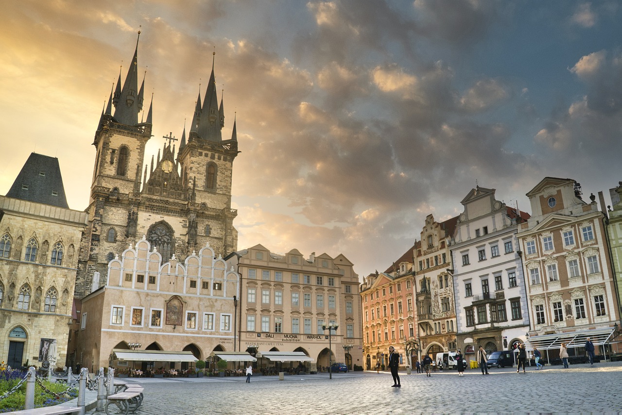 Historischer Platz mit gotischer Kirche und umliegenden barocken Gebäuden, wenige Menschen auf dem Kopfsteinpflaster bei Sonnenuntergang.