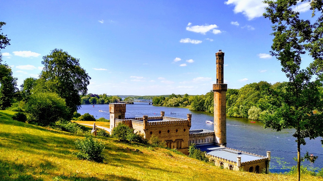 Orientalisch anmutendes Bauwerk mit hohem Turm am Wasser, umgeben von Bäumen und mit Brücke im Hintergrund.
