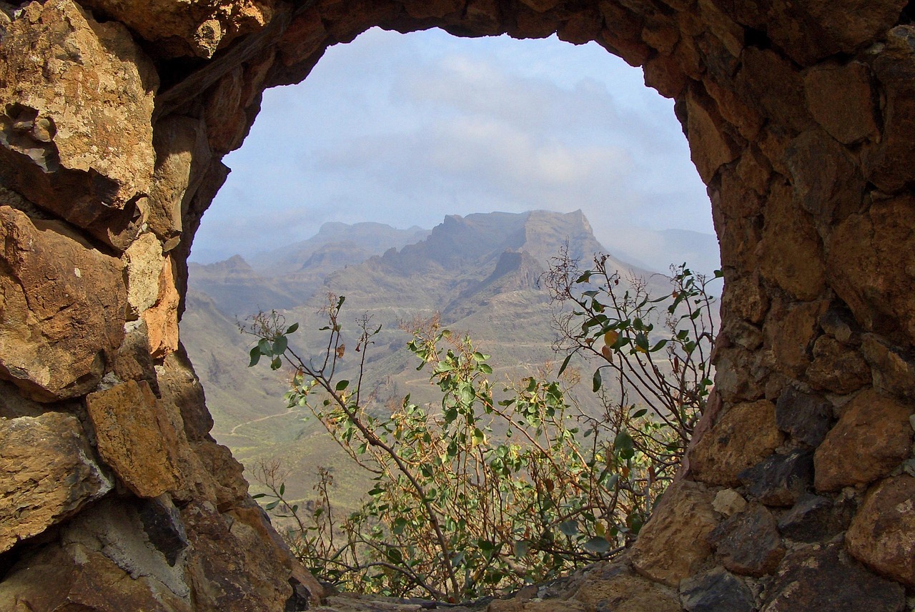 Blick durch ein rundes Felsenfenster auf ein Gebirgspanorama mit spärlicher Vegetation im Vordergrund.