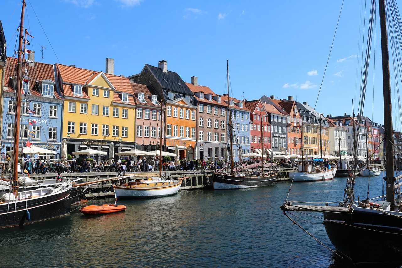 Bunte Häuserreihe mit roten Dächern an einer Uferpromenade, davor Segelboote im Wasser.