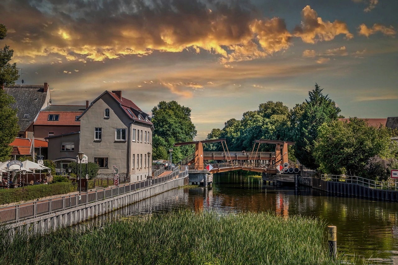 Stadtansicht mit Fußgängerbrücke über Kanal, Häusern am Ufer und dramatisch beleuchtetem Abendhimmel.