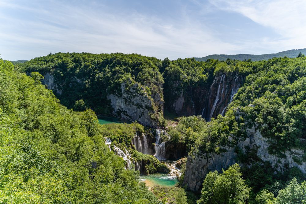 Mehrere Wasserfälle stürzen in türkisfarbene Becken, umgeben von dicht bewaldeten Felswänden in hügeliger Landschaft.