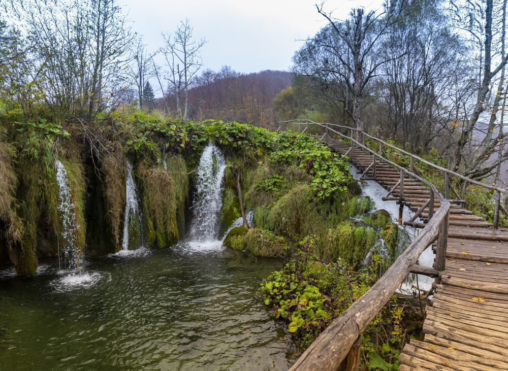Mehrere kleine Wasserfälle fließen über moosbewachsene Felsen in ein Becken, daneben ein hölzerner Steg mit Geländer in bewaldeter Umgebung.