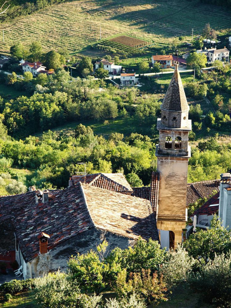 Kirchturm mit spitzem Dach ragt über rote Ziegeldächer eines Dorfes, im Hintergrund Felder, Bäume und verstreute Häuser.