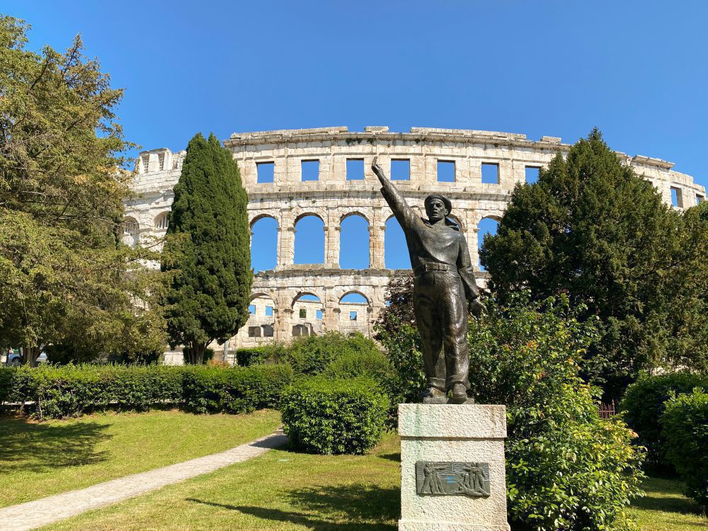 Bronzestatue eines Mannes mit erhobenem Arm auf Sockel, im Hintergrund römisches Amphitheater, umgeben von Bäumen und Rasenfläche.