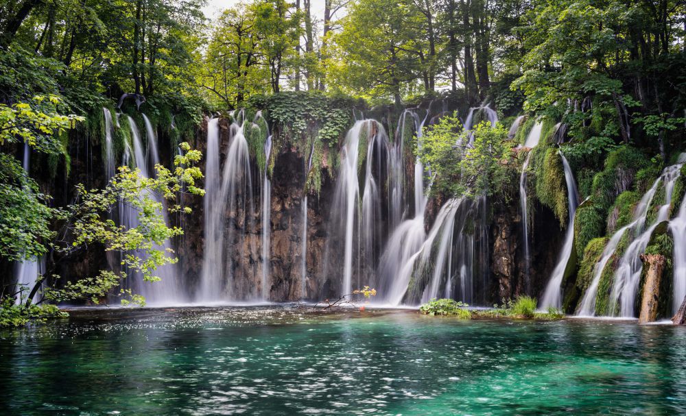 Breiter Wasserfall mit mehreren Kaskaden, die zwischen Felsen und grünem Bewuchs in ein türkisfarbenes Becken stürzen.