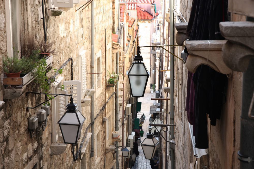 Enge Gasse mit Kopfsteinpflaster, Laternen, Fensterläden, Pflanzenkästen und Wäscheleinen zwischen historischen Steinfassaden.