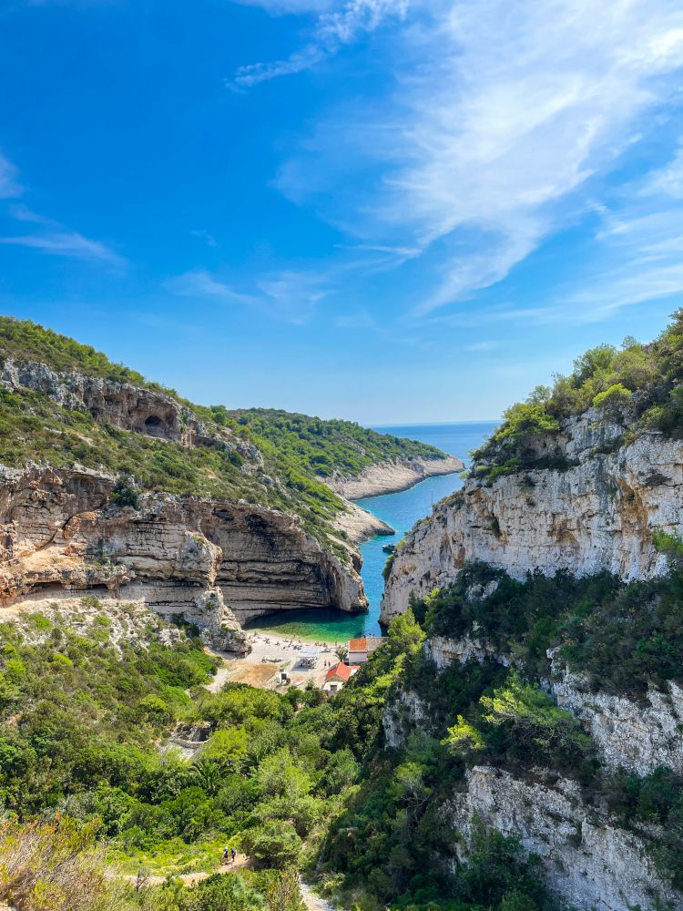 Bucht mit türkisfarbenem Wasser, Kiesstrand und wenigen Gebäuden, eingerahmt von steilen, bewaldeten Felsen unter blauem Himmel.
